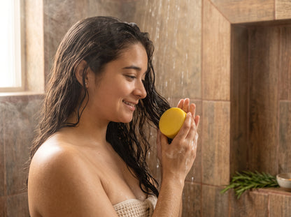 Mujer de cabello largo y mojado, sonriendo en la ducha, con una barra de acondicionador sólido de romero y cúrcuma en la mano
