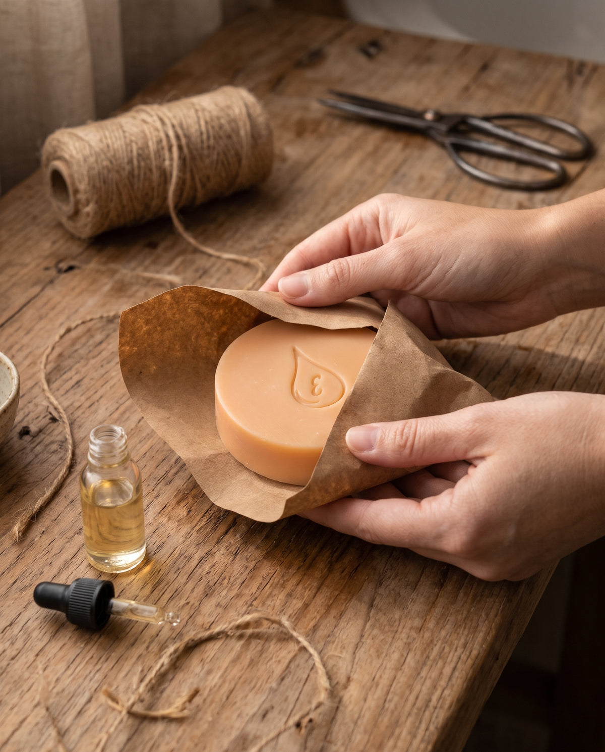 Manos envolviendo un jabón artesanal de Vitamina E en papel kraft sobre una mesa de madera, con hilo de yute, tijeras y un frasco con gotero de aceite