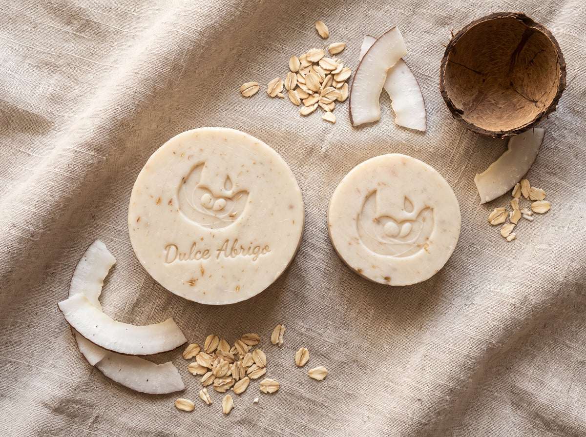 Jabón artesanal de avena y coco Dulce Abrigo en barra de 110 g junto a la barra pequeña de 50 g, sobre lino con copos de avena, trozos de coco y media cáscara de coco