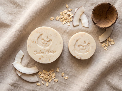 Jabón artesanal de avena y coco Dulce Abrigo en barra de 110 g junto a la barra pequeña de 50 g, sobre lino con copos de avena, trozos de coco y media cáscara de coco