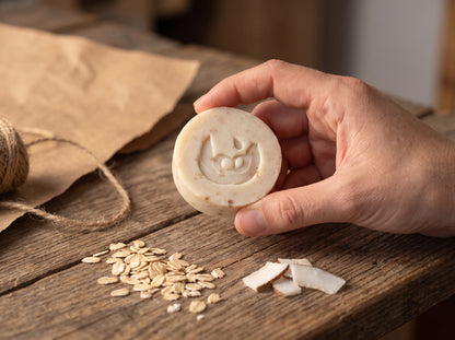 Mano sosteniendo la barra pequeña de 50 g del jabón artesanal de avena y coco, con el sello grabado, junto a copos de avena, trozos de coco y papel kraft con hilo de yute