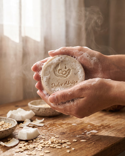 Manos haciendo espuma con un jabón artesanal de avena y coco frente a una ventana con luz suave y vapor